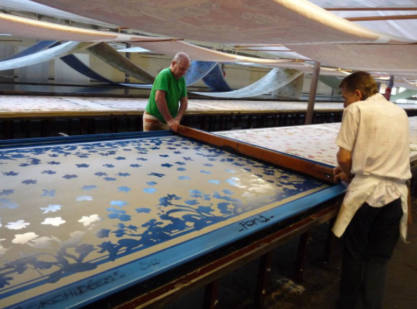 Two printers carefully lift up a printing screen to place it one the fabric. Using a squeegee, color is spread evenly across the screen onto the cloth. Each application of color requires the use of a new screen.